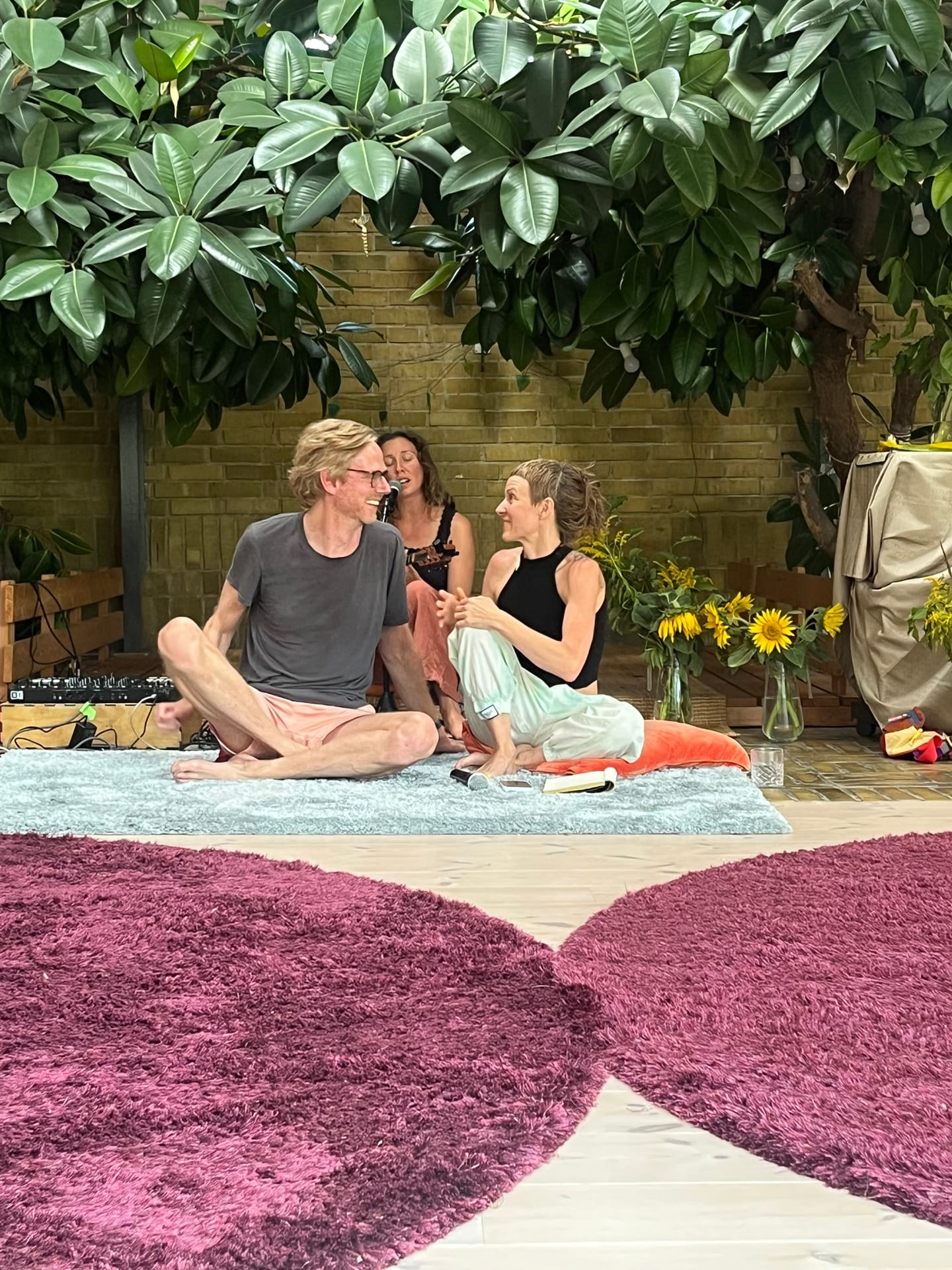 Two people sitting on a rug in the Pool Room, laughing together, with sunflowers behind them